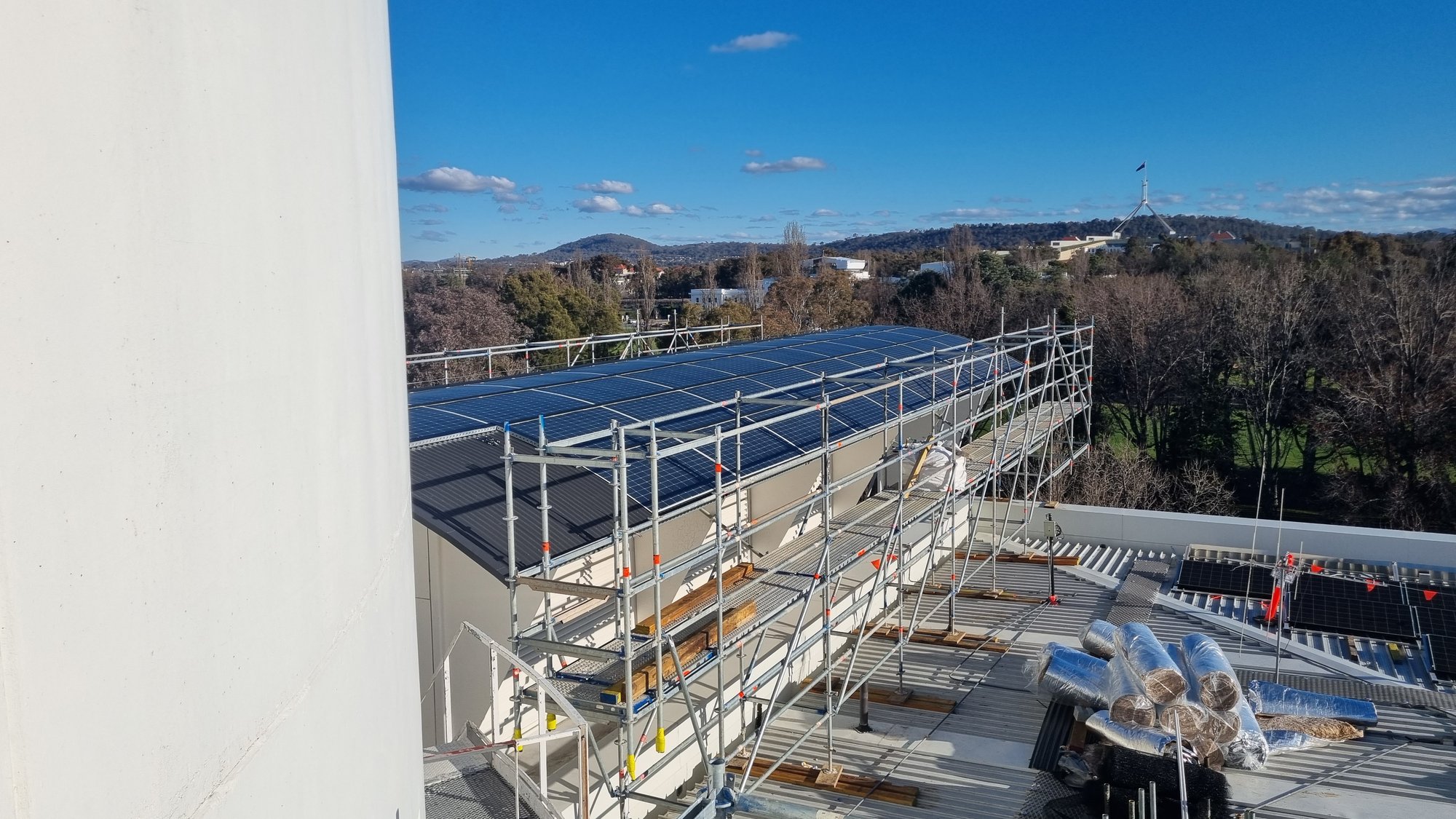 Curved rooftop solar array under scaffold, with the Parliament House flagpole on the Canberra horizon