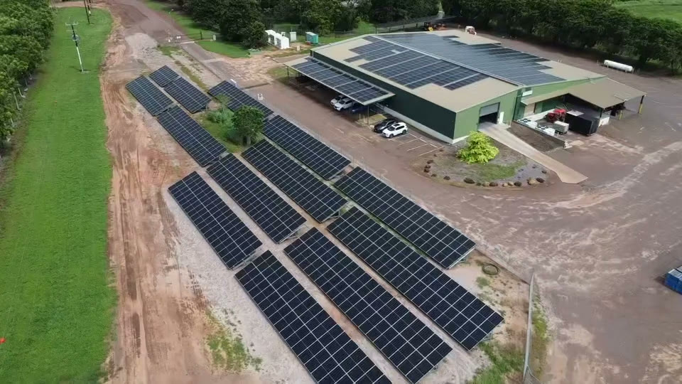 Aerial of a Northern Territory agricultural site: rows of ground mount solar on red earth beside a green shed carrying a full rooftop array and a carport